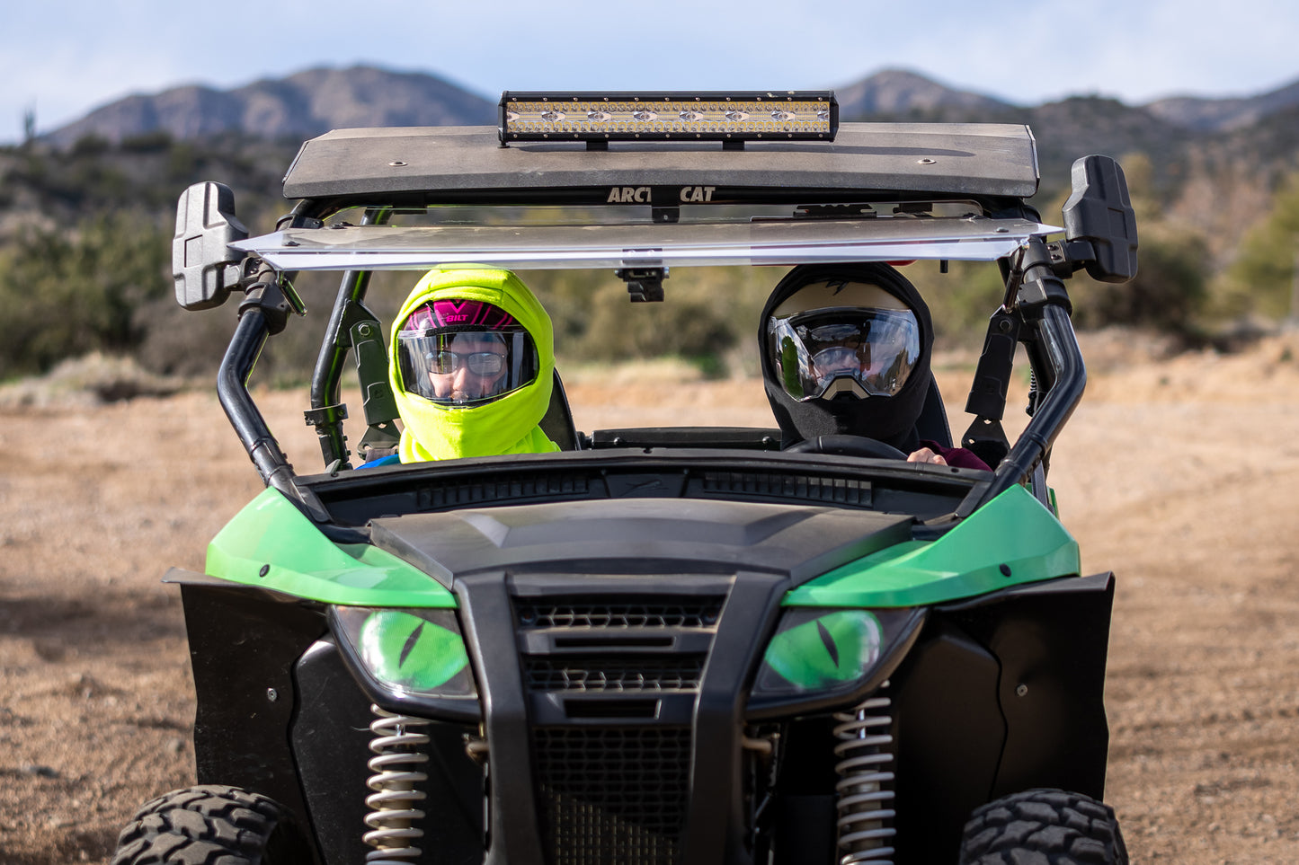 Two people using green and black helmet covers on side by side all terrain vehicle for dust filtration in desert